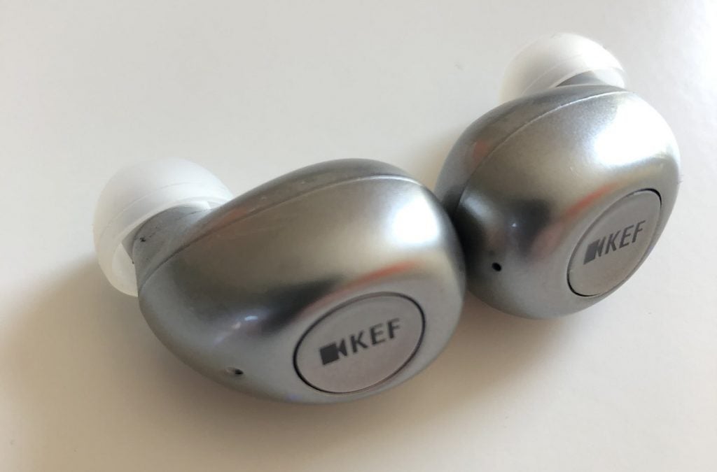 Silver and white KEF Mu3 earbuds resting on white background, back left top view, a video camera logo with KEF printed in the back