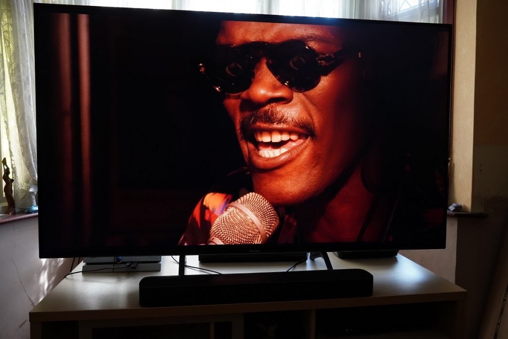 A black Panasonic TX 65HX940B TV standing on a wooden table displaying a man wearing glasses singing, a scene from Do the right thing