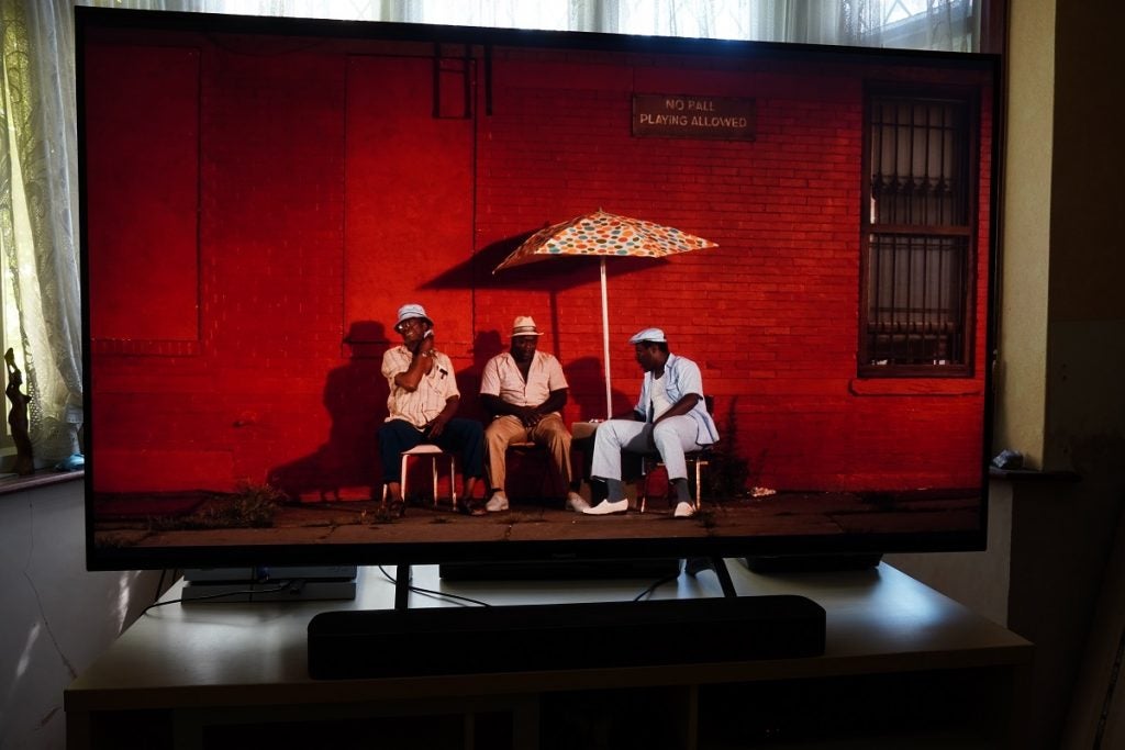 A black Panasonic TX 65HX940B TV standing on a wooden table displaying 3 men sitting on chairs under an unmbrella against a red wall, a scene from Do the right thing