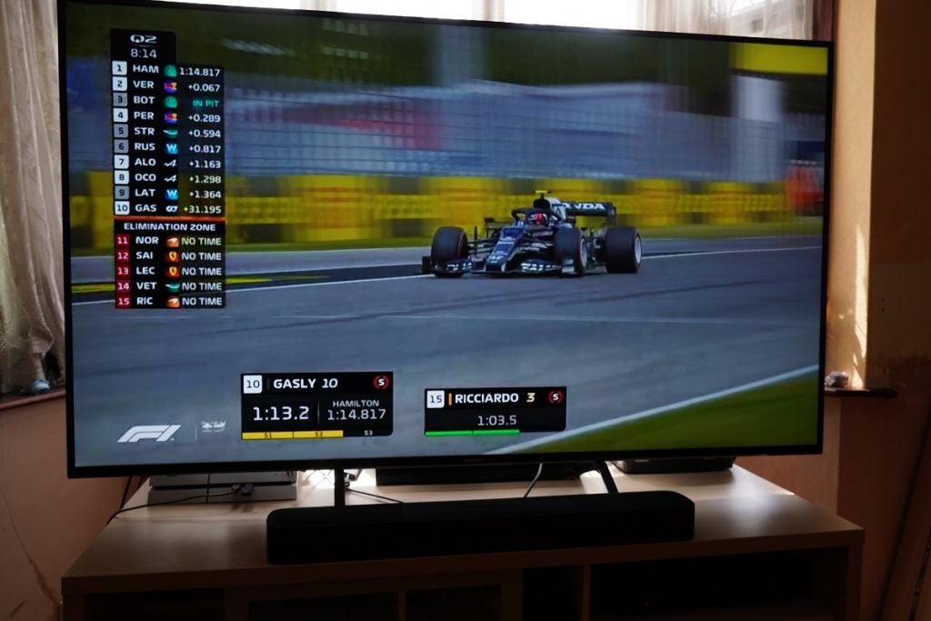A black Panasonic TX 65HX940B TV standing on a wooden table displaying F1 race with a list of racers on the left