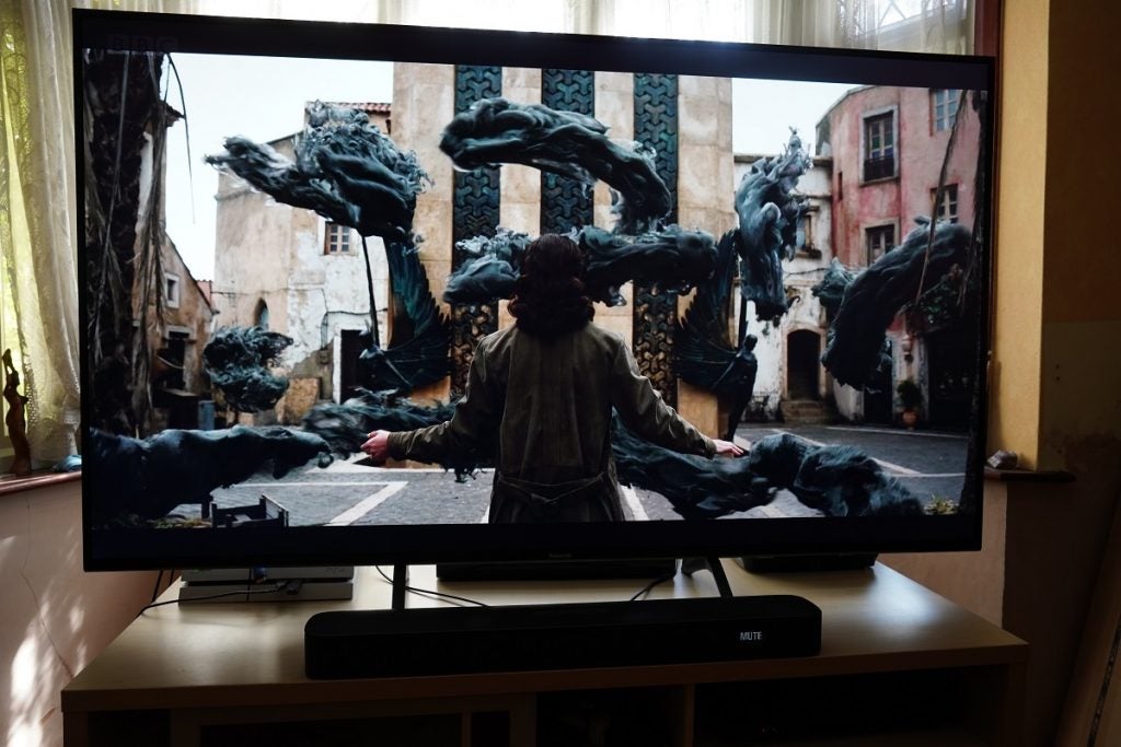 A black Panasonic TX 65HX940B TV standing on a wooden table displaying a scene from His Dark Materials 2, a man sorrounded by black smoke, showcasing black magic