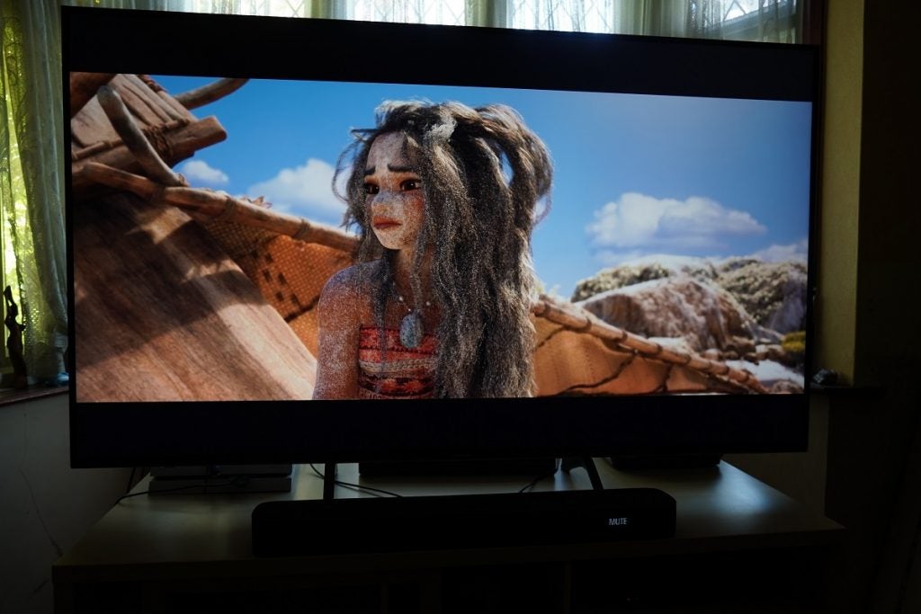 A black Panasonic TX 65HX940B TV standing on a wooden table displaying a scene from Moana, Moana covered in sand