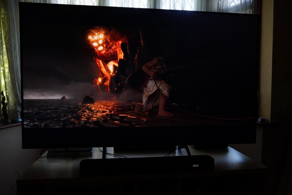 A black Panasonic TX 65HX940B TV standing on a wooden table displaying a scene from Moana, Moana handling a boat and a fire monster looking at her
