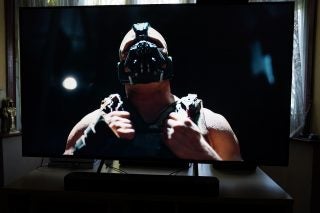 A black Panasonic TX 65HX940B TV standing on a wooden table displaying a close up image of man wearing a weird face mask