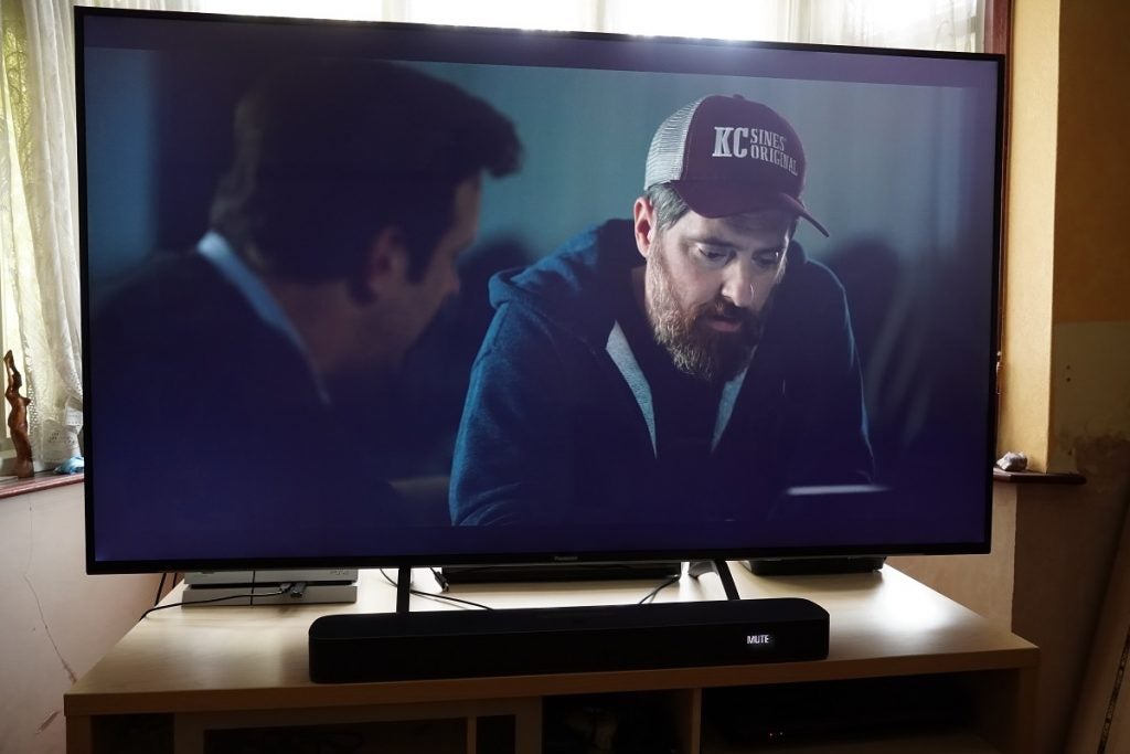 A black Panasonic TX 65HX940B TV standing on a wooden table displaying two men sitting, one wearing sports outfit with cap and the other in a black suit