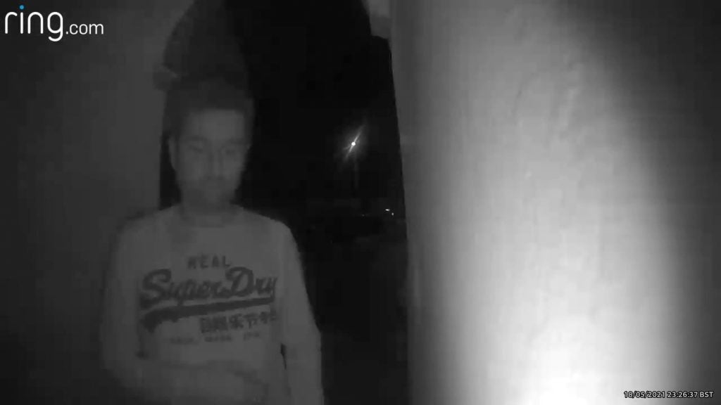 Ring Video Doorbell Wired night sampleA man in a red t-shirt, standing on the front door at night, looking towards the camera of Ring video  doorbell, showcasing night sample with floodlight turned on, black and white picture