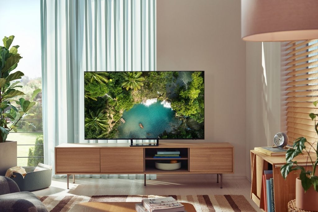 Image of well furnished room with a Samsung AU9000 TV standing on small hieghted shelf, displyaing a boat in a river sorrounded by trees, view from top