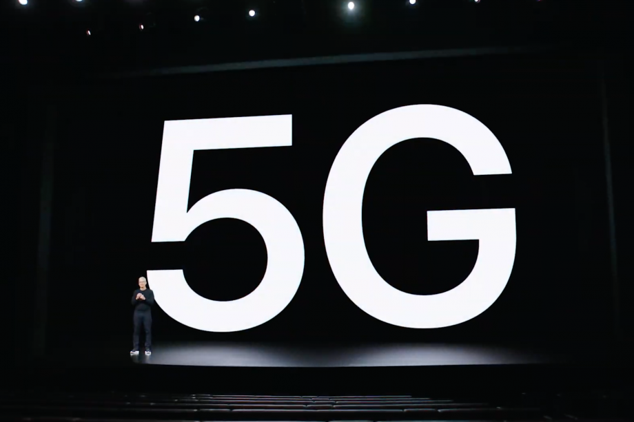 Tim Cook in a black outfit standing on black stage with black chairs on front with 5G displayed in white on black background on a screen behind him