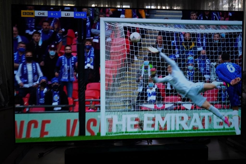 LG G1 OLED TV playing football, displaying a goalkeeper jumping to stop football which is abou to reach goal net, audience majorly in blue outfit sitting behind gaol post