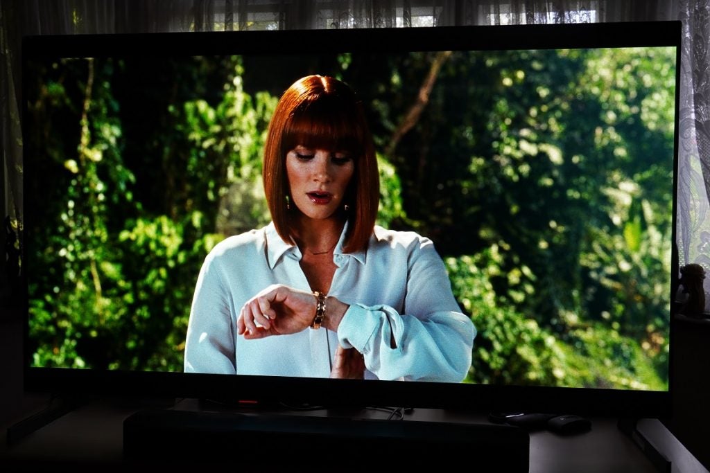 LG G1 OLED TV playing Jurrasic World, displaying  a women wearing white outfit looking at her watch in left hand and trees behind