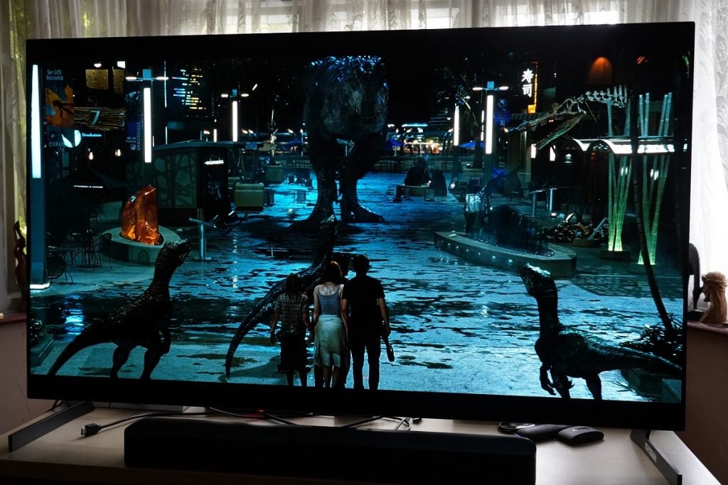 LG G1 OLED TV playing Jurrasic World, displaying  a women and two boys standing behind three small dinosaurs and a huge dinosaur approching them 