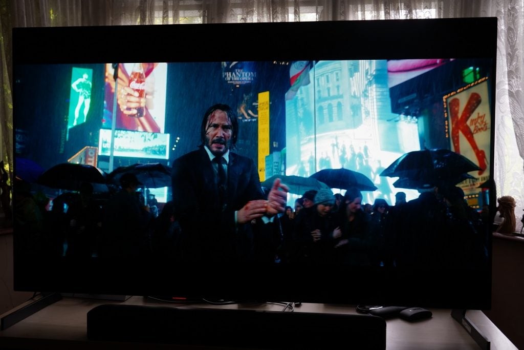 John Wick 3 on the LG G1 OLEDLG G1 OLED TV playing a movie, displaying night scene, a man wearing a coat, standing in rain, other people going around, big screens advertising behind