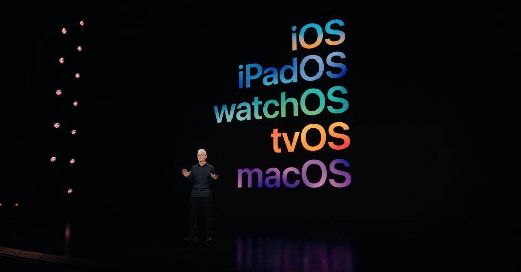 Tim cook standing on stage in a black outfit with iOS, iPadOS, watchOS, tvOS, and macOS written on the screen behind him