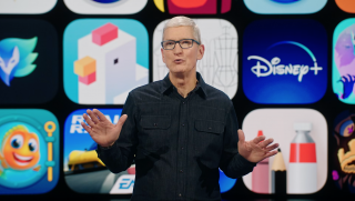 Tim Cook standing on a stage in a black outfit with multiple icons displayed on a big screen behind him