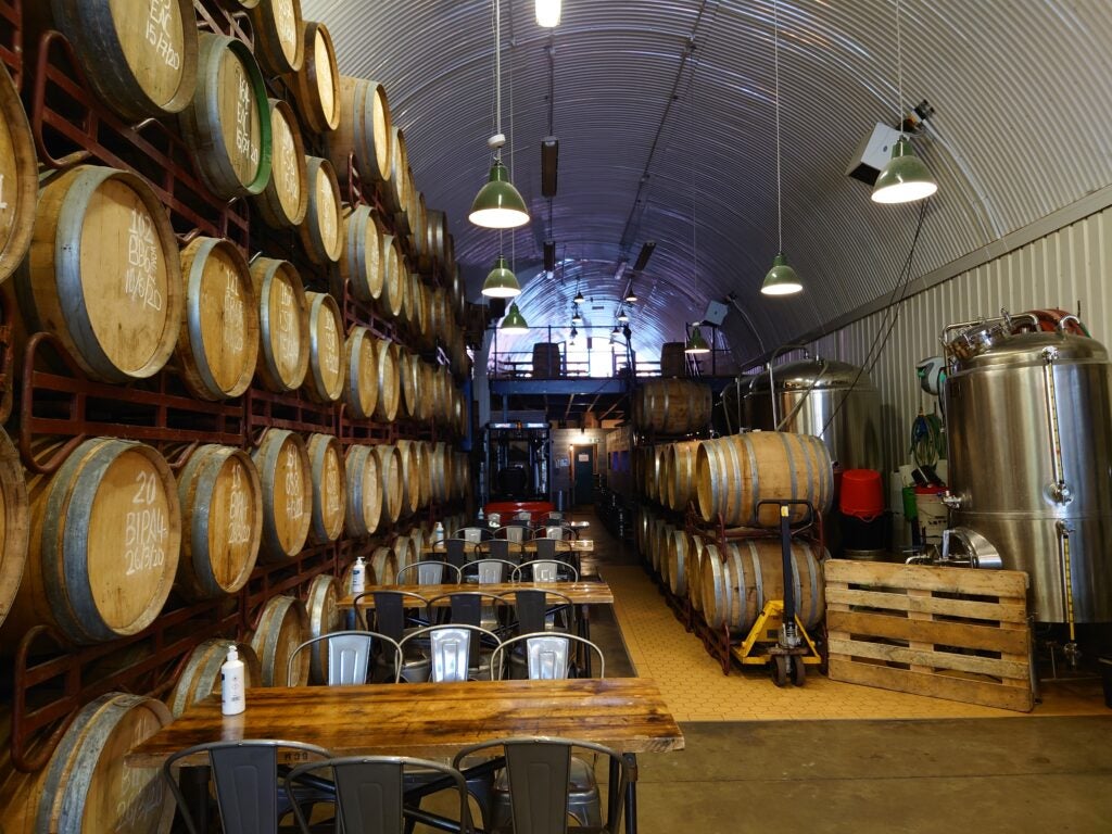 Interior of a brewery with stacked wooden barrels and equipment.