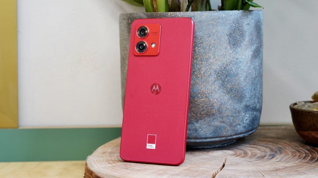 Moto G84 5G against a plant potRed Motorola Moto G84 5G smartphone with dual cameras on wood.
