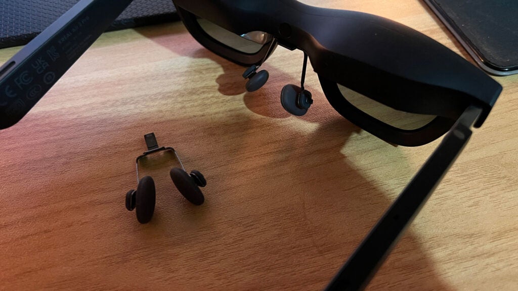 A close-up of the Xreal Air 2 Pro glasses with an attached nose pad and a different, smaller nose pad on the desk.Xreal Air 2 Pro smart glasses and charging clip on desk.